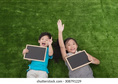 Cute Asian Children Lying And Holding Blackboard On Green Grass