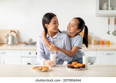 Cute Asian Daughter Hugging Her Young Happy Mom While Enjoying Homemade Cookies And Biscuits Together At Home, Sitting In Kitchen Interior. Family Lifestyle, Motherhood