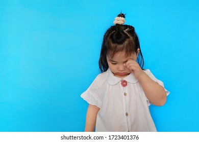 A Cute Asian Female Toddler Feeling Upset, Crying And Wiping Tear From Her Eye With Her Hand. Plain Blue Background.  