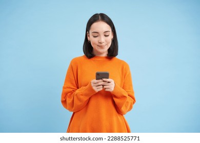 Cute asian girl looks at her cellphone and smiles, uses mobile application, social media app, playing game on phone, standing over blue background.