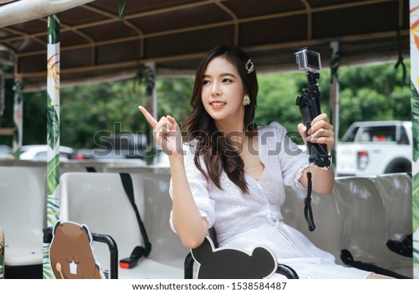 Cute asian girl on white dress sitting on\
shuttle bus holding stick action\
camera.