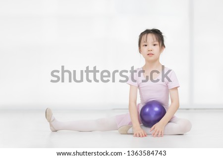 Cute Asian girl in pink dress is posing and holding purple gymnastic ball in front of window.