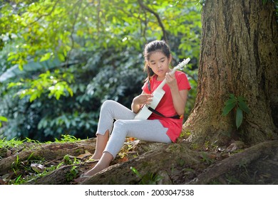 Cute Asian Girl Playing Ukulele Under Tree.Pretty Child With Musical Instrument In Outdoor.Young Female Spending Leisure Time With Ukelele In Park During Summer.Music Activity For Student.