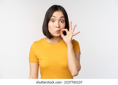 Cute asian girl seal lips, zipping mouth with finger, promise to keep secret, taboo gesture, standing in yellow tshirt over white background