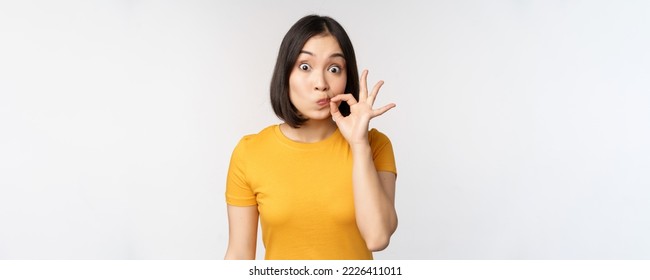 Cute asian girl seal lips, zipping mouth with finger, promise to keep secret, taboo gesture, standing in yellow tshirt over white background. - Shutterstock ID 2226411011