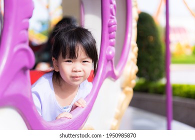 Cute Asian Girl Sitting In Carousel Ride. Happy Child Sit And Watch From Window Of The Player. Kid Smiles Sweetly. Children Squint Due To The Sunlight. Baby Wearing A White Dress Is 3-4 Years Old.