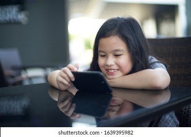 Cute Asian Girl Smiling Holding Smart Phone On The Table.Child Using Gadget In Restaurant.Modern Kid Watching Wifi Movie Online.Young Female With Device.Screen Game Time.Internet Line For Children.