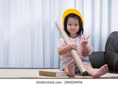 Cute Asian girl wearing a yellow toy hat like an engineer, she holds a roll of brown paper tube, a big playbook next to the little kid. Child role play to be at home or school.