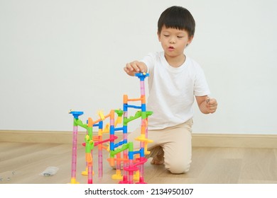 Cute Asian Kindergarten Boy Child Sitting On Floor Having Fun Playing With Colorful Plastic Marble Run Track Toy Set, Activities At Home, Educational And Fun Toys For Young Kid, Isolated On White Wall