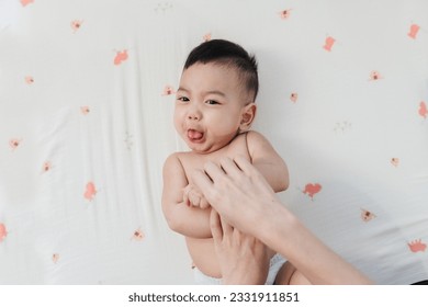 Cute Asian little boy in good mood sticks tongue out while Mother’s Finger Applying Skin Care Lotion on Her Son’s Arm in Closeup Shot, Embracing the Concept of Baby Care.