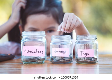 Cute Asian Little Child Girl Putting Coin Into Glass Bottle In The Garden. Kid Saving Money For The Future Concept