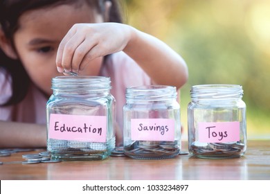 Cute Asian Little Child Girl Putting Coin Into Glass Bottle In The Garden. Kid Saving Money For The Future Concept