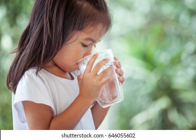 Cute Asian Little Child Girl Drinking Fresh Water From Glass On Green Nature Background