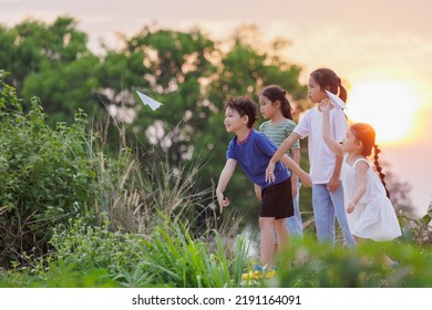 Cute Asian Little Girl And Her Siblings Playing With Paper Airplane Toy Together With Fun. Group Of Asian Kid Throwing Paper Plane And Playing Together In The Countryside At The Sunset Time.