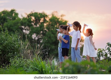 Cute Asian Little Girl And Her Siblings Playing With Paper Airplane Toy Together With Fun. Group Of Asian Kid Throwing Paper Plane And Playing Together In The Countryside At The Sunset Time.