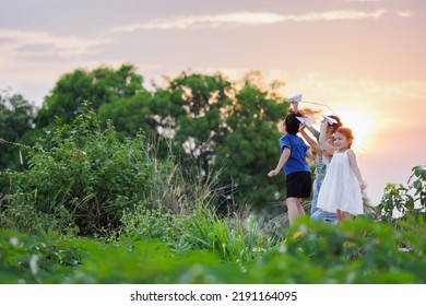 Cute Asian Little Girl And Her Siblings Playing With Paper Airplane Toy Together With Fun. Group Of Asian Kid Throwing Paper Plane And Playing Together In The Countryside At The Sunset Time.