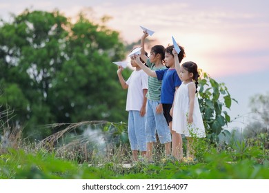 Cute Asian Little Girl And Her Siblings Playing With Paper Airplane Toy Together With Fun. Group Of Asian Kid Throwing Paper Plane And Playing Together In The Countryside At The Sunset Time.