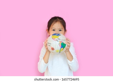 Cute Asian Little Kid Girl Holding A Globe With Looking At Camera Isolated On Pink Background. Portrait Of Child With The Earth Globe Sphere. Focus At Children Face.