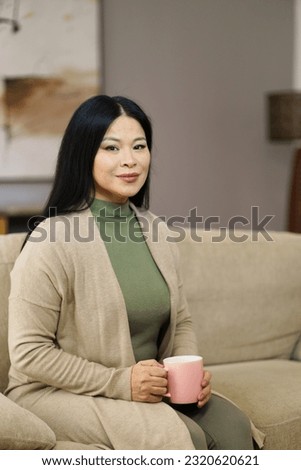Cute Asian MILF sitting on couch with cup of coffee home. The image captures moment of relaxation and leisure as woman enjoys her coffee break in comfort of her own home. . High quality photo