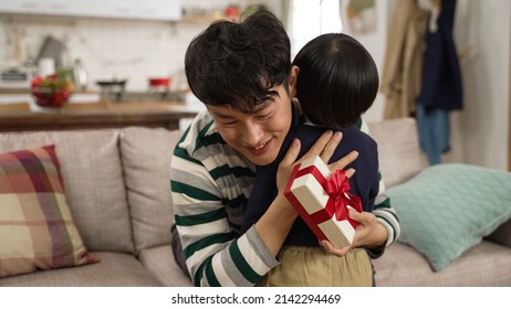 Cute Asian Preschool Boy Surprising Father With A Gift On Father’s Day. Smiling Asian Dad Feeling Touched And Hugging His Baby Son In The Living Room At Home