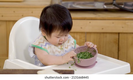 Cute Asian Toddler Wearing A Bib Is Looking Curiously At The Messy Table On The Baby Chair While Playing With The Bowl Of Vegetable Porridge.