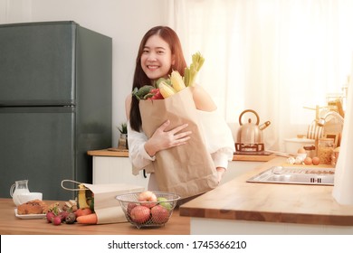 Cute Asian Woman Holding Shopping Paper Bag With Fruits And Vegetables Stand In The Kitchen,her Look At Camera With Brightly Smile.