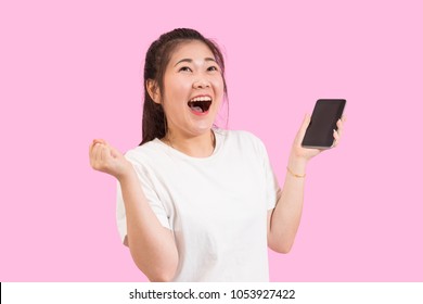 Cute Asian Woman Holding Smart Phone In Hand, Screams With Joy And Happiness, Suprise Feeling, Winner Expression, White Shirt, Pink Background