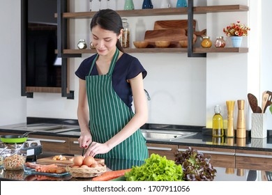 Cute Asian Woman With Long Hair Wearing A Green Apron Cooking In The Kitchen. In Her House On Holiday, On The Table There Is A Gas Stove, Eggs, Vegetables And Salads To Cook For Breakfast.Smiling Face