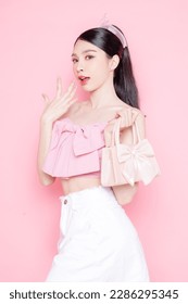 Cute Asian woman model gathered in ponytail with korean makeup style on face have plump lips and clean fresh skin wearing pink camisole holding handbag on isolated pink background. - Shutterstock ID 2286295345