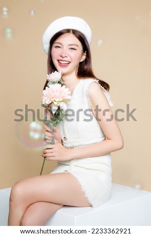 Cute Asian woman with perfect skin and holding pastel flower. Pretty girl model wearing white beret and natural makeup on beige background. Cosmetology, beauty and spa, wellness, Plastic surgery.