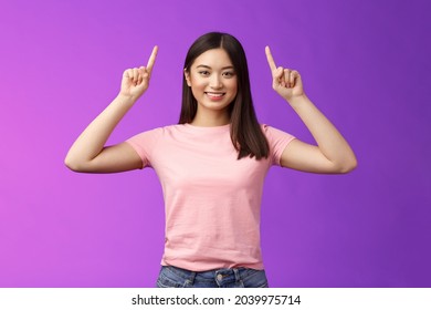 Cute assertive dark-haired asian woman wear pink t-shirt raise hands up, showing promo, pointing fingers up, grinning, inviting use link, try-out product, give direction, stand purple background - Powered by Shutterstock