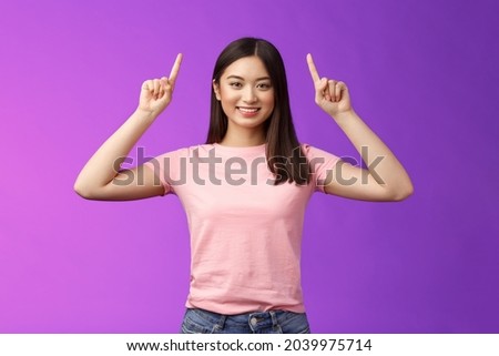Cute assertive dark-haired asian woman wear pink t-shirt raise hands up, showing promo, pointing fingers up, grinning, inviting use link, try-out product, give direction, stand purple background