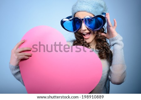 Cute attractive fashion young girl posing with funny big love glasses and pink heart, over blue background