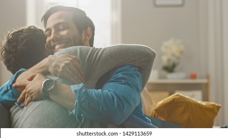 Cute Attractive Male Gay Couple Sit Together On A Sofa At Home. Boyfriends Are Hugging And Embracing Each Other. They Are Joyful And Laughing. They Are Casually Dressed And Room Has Modern Interior.