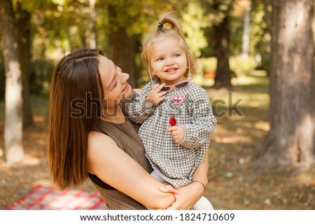 Cute attractive photo of a young woman hugging her baby. A beautiful girl looks at her daughter and smiles. A little girl is holding a lollipop in her hands. Stand together in the park. 