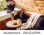 Cute autumn hygge home decor arrangement. Tiny wooden cabin balcony with heather flowers, lavender in bottle vase, candlelight flame, soft beige plaid waiting on comfortable garden furniture chair.