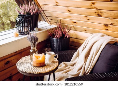 Cute Autumn Hygge Home Decor Arrangement. Tiny Wooden Cabin Balcony With Heather Flowers, Lavender In Bottle Vase, Candlelight Flame, Soft Beige Plaid Waiting On Comfortable Garden Furniture Chair.