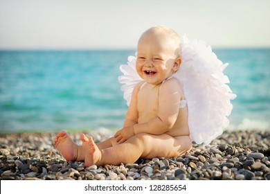 Cute Baby With Angel Wings Is Sitting On The Seashore