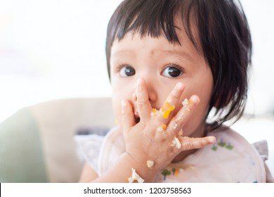 Cute Baby Asian Child Girl Eating Healthy Food By Herself And Making A Mess On Her Face And Hand