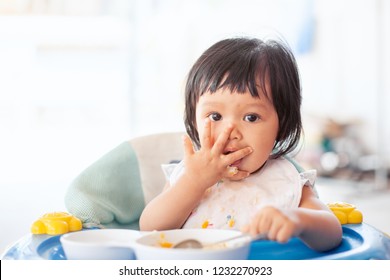 Cute Baby Asian Child Girl Eating Healthy Food By Herself And Making A Mess On Her Face And Hand