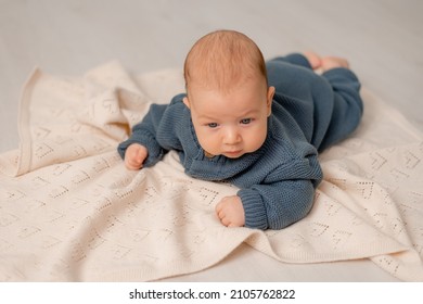 Cute Baby With Blue Eyes In Blue Knitted Jumpsuit Lies On His Tummy On Gray Plaid. Child Learns To Crawl. Motherhood And Childhood. Solid Background. Space For Text. High Quality Photo