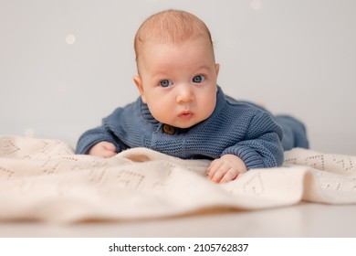 Cute Baby With Blue Eyes In Blue Knitted Jumpsuit Lies On His Tummy On Gray Plaid. Child Learns To Crawl. Motherhood And Childhood. Solid Background. Space For Text. High Quality Photo