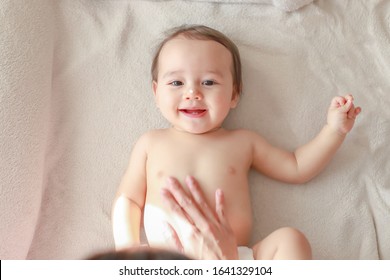 Cute Baby Boy Applying Lotion Cream On Face And Body By His Mommy Making Massage After Bathing. Newborn Child Temporary First Milk Teeth Smiling Face Top View. Mixed Race Asian-German Kid.