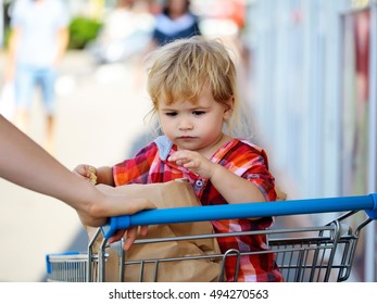 Cute Baby Boy Blond Child In Plaid Shirt Sits In Shopping Trolley And Eats Cookie From Paper Packet Outdoors On Sunny Day