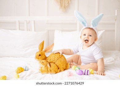 cute baby boy with bunny ears and colorful eggs on a white bed at home playing, little blonde baby with a rabbit, happy easter concept - Powered by Shutterstock