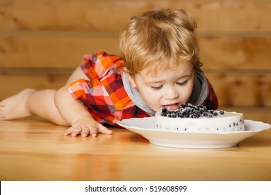 Cute baby boy child with blond curly hair eats delicious cake with blueberries lying on wooden table