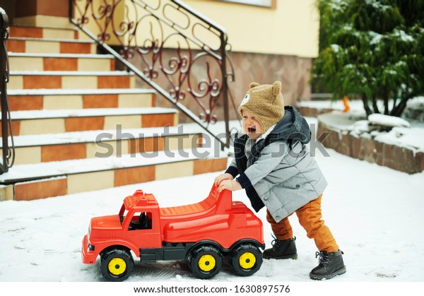 Cute baby\
boy in cozy winter clothes and funny hat making his first steps\
with toy car in snow on beauty winter\
day