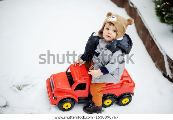 Cute baby boy in cozy winter
clothes and funny hat playing with toy car in snow on beauty winter
day