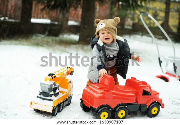 Cute baby boy in cozy winter clothes and funny hat\
playing with toy cars truck and lifting crane machine in snow on\
beauty winter day