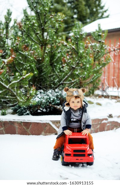 Cute baby boy in cozy winter\
clothes and funny hat playing with toy car in snow on beauty winter\
day